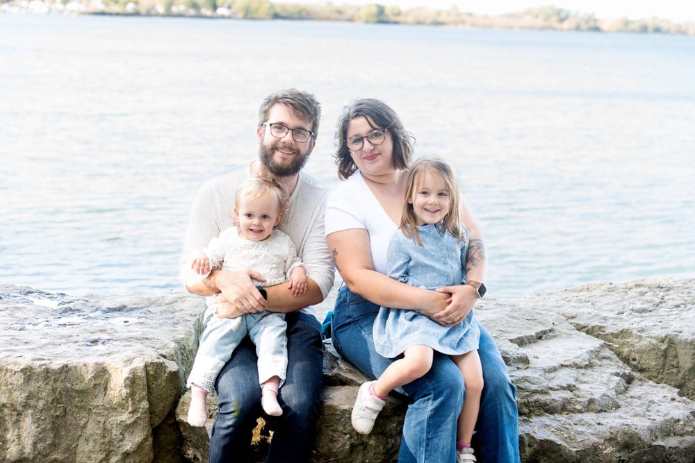 Family sitting together on rocky shoreline by water during outdoor photo session.