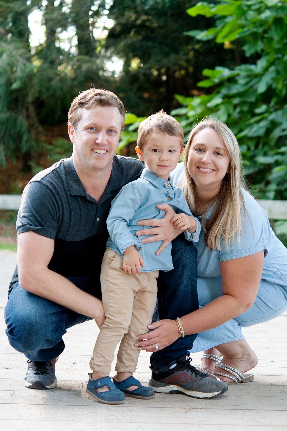 Family of three pose together outdoors on a sunny day surrounded by greenery.