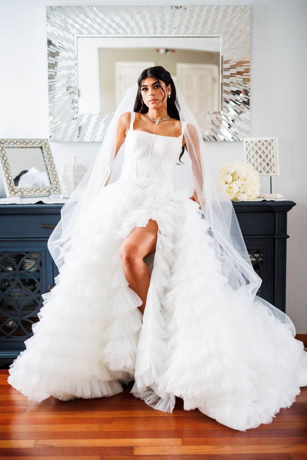 A bride in a luxurious white ruffled ballgown poses elegantly in front of a decorative mirror.