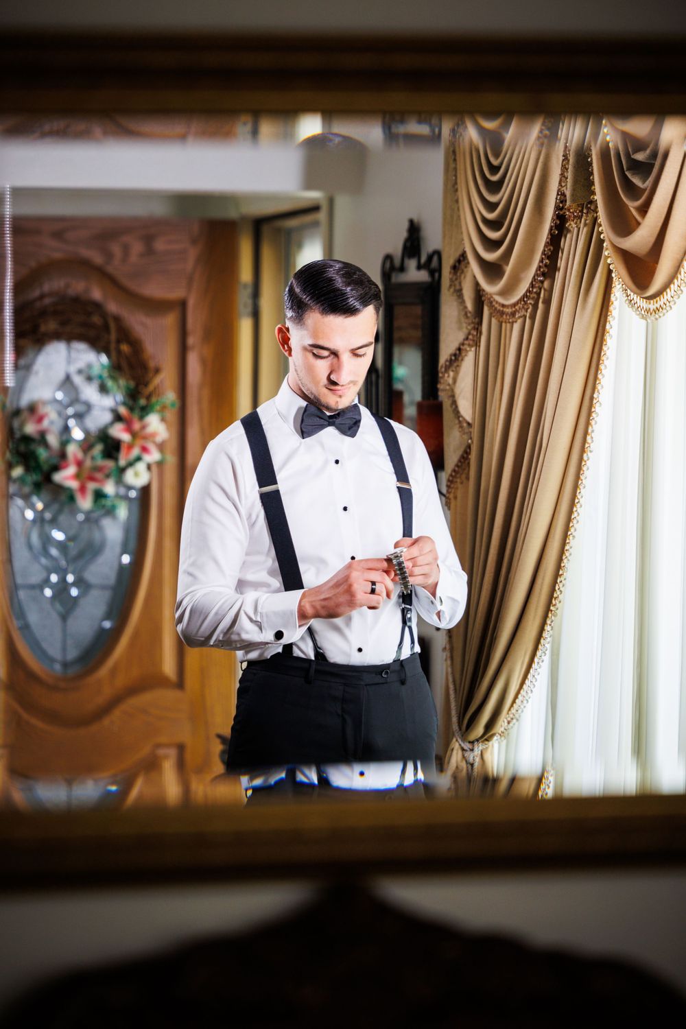 A groom in classic white shirt and suspenders adjusts his cuffs while getting ready.