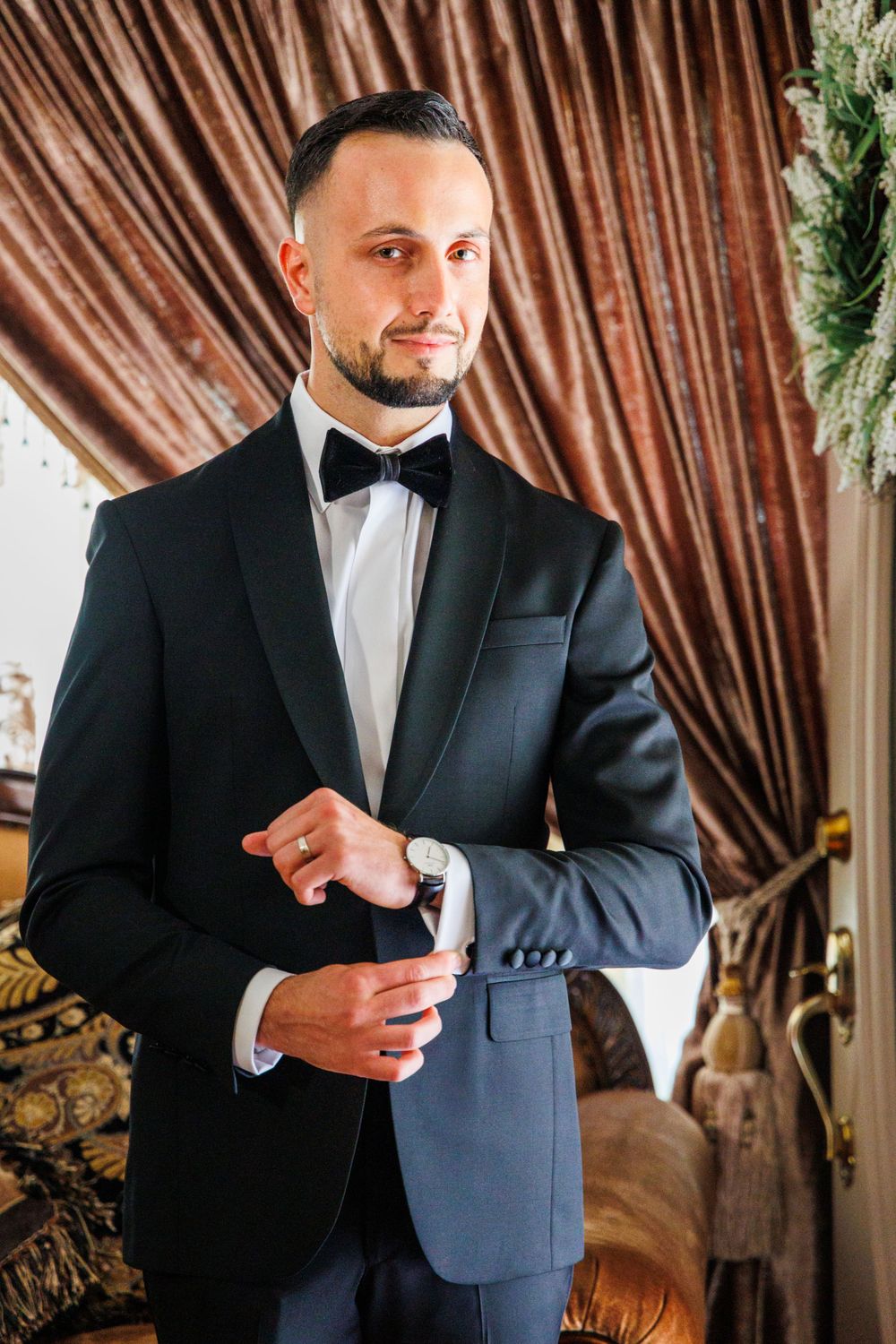 Groom adjusting cufflinks while wearing classic black tuxedo against textured backdrop.