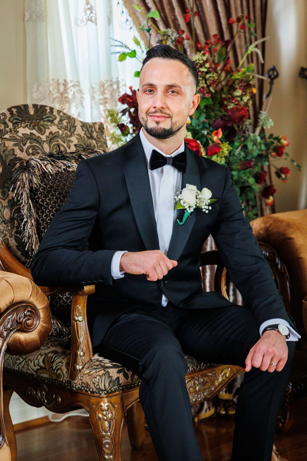 Groom in classic black tuxedo seated in ornate vintage chair with floral arrangements.