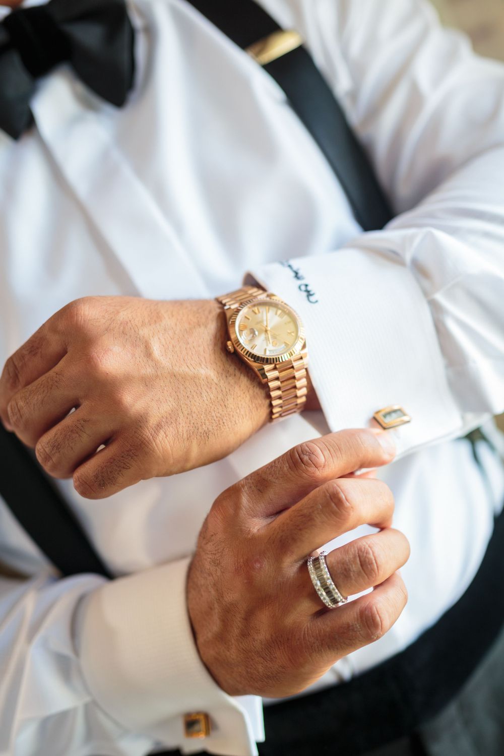 Close up of groom's hands showing gold watch, cufflinks and ring details with bow tie visible.