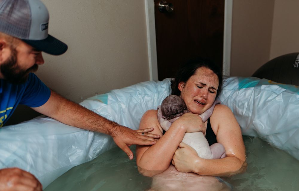 woman in a birth pool, holding her just birthed baby on her chest, hugging tightly and crying, while dad touches mom