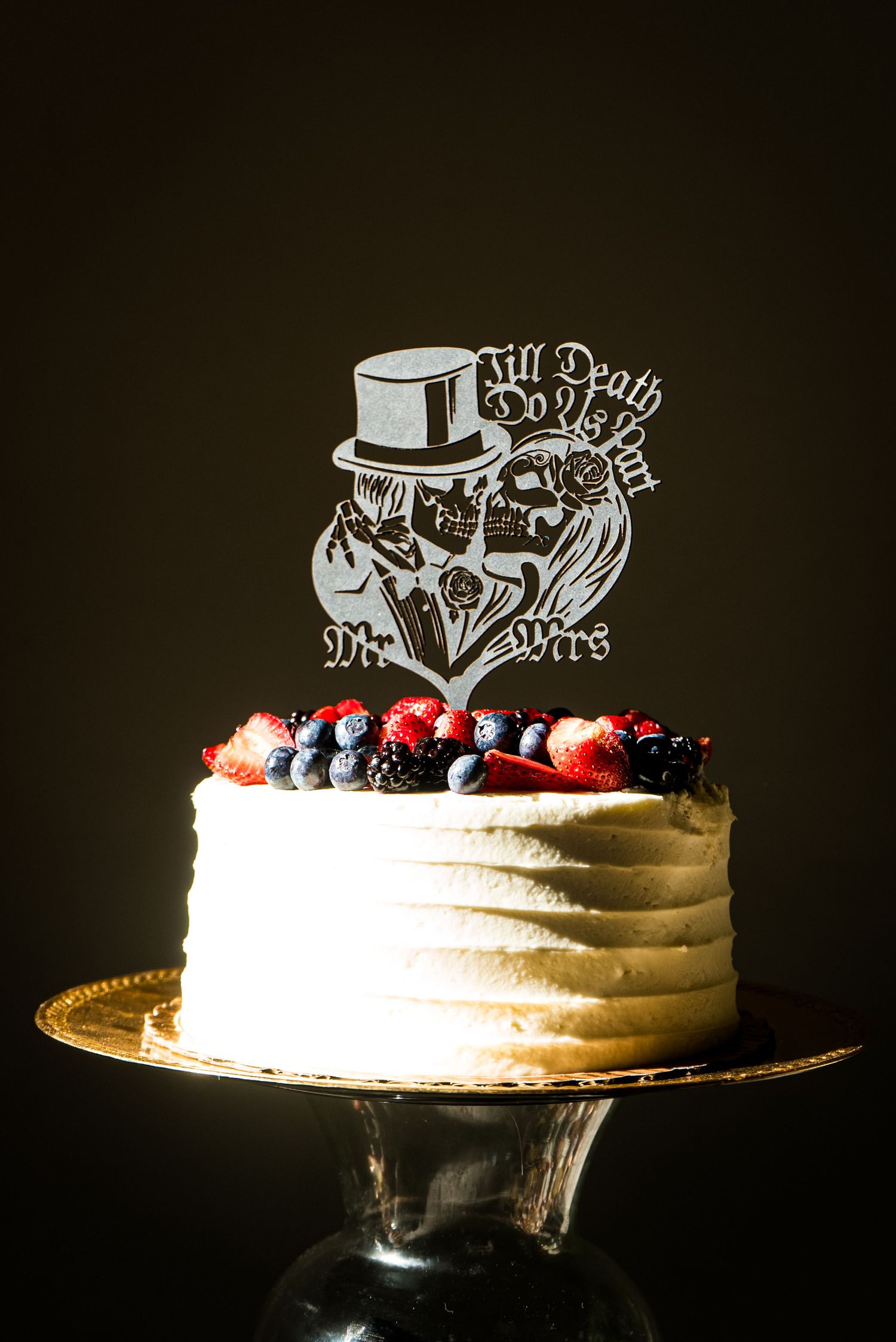 Wedding cake with 'Till Death Do Us Part' topper decorated with fresh berries on a cake stand.