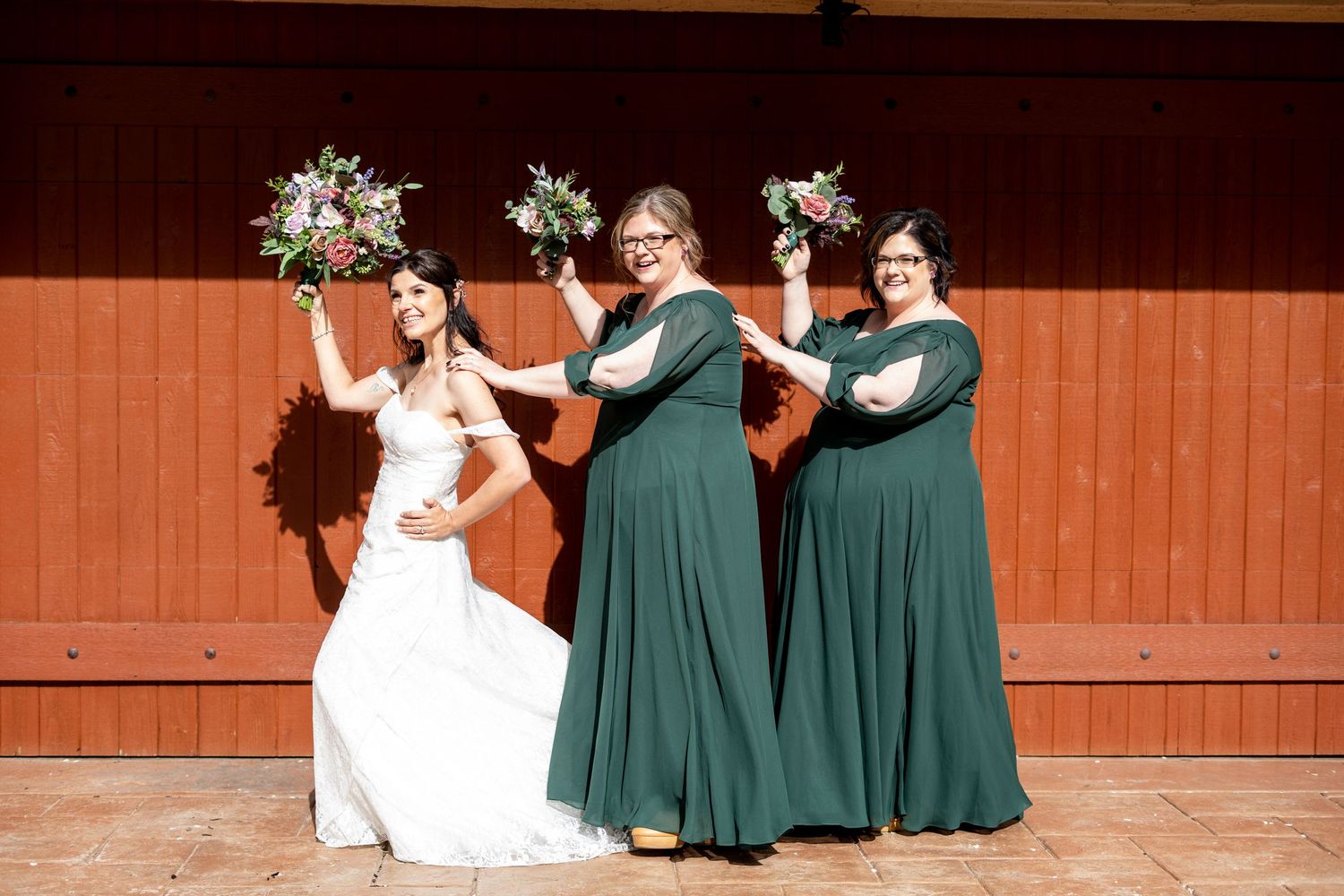Bride and bridesmaids in green dresses pose playfully with bouquets against a rustic red wall.