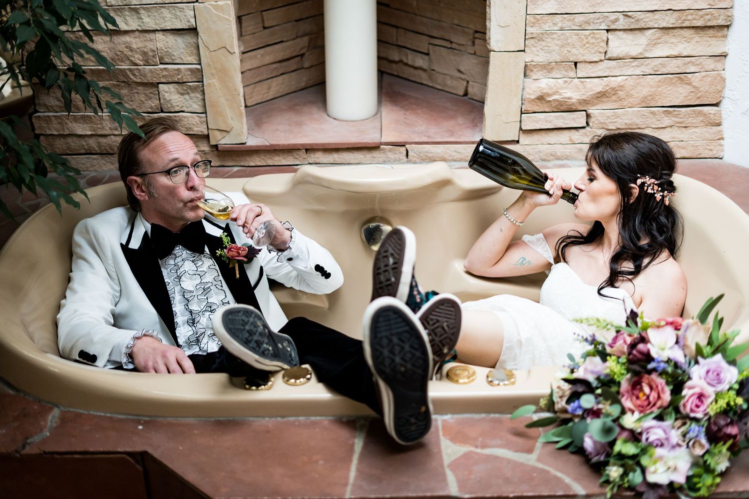 Groom and bride playfully enjoying drinks while lounging in tub with wedding bouquet nearby.