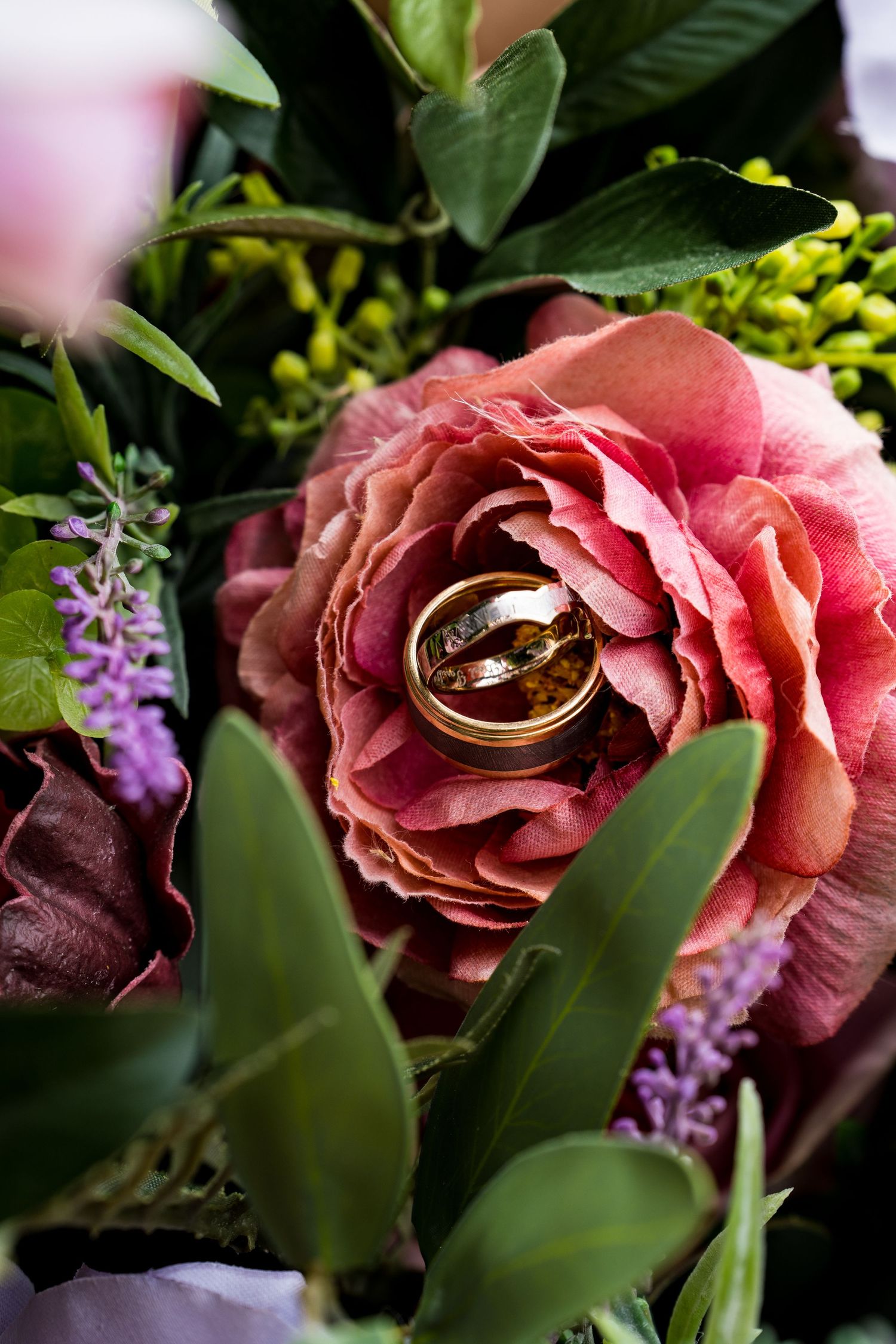 Wedding rings nestled in a pink rose bloom surrounded by purple flowers and greenery.