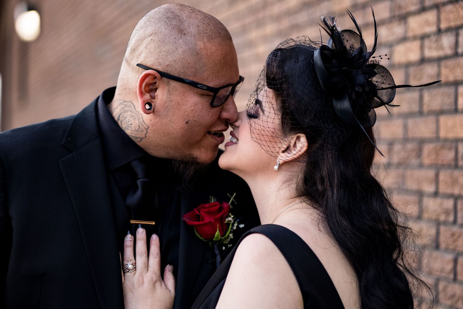 A couple in formal attire share a romantic kiss against a brick wall background.