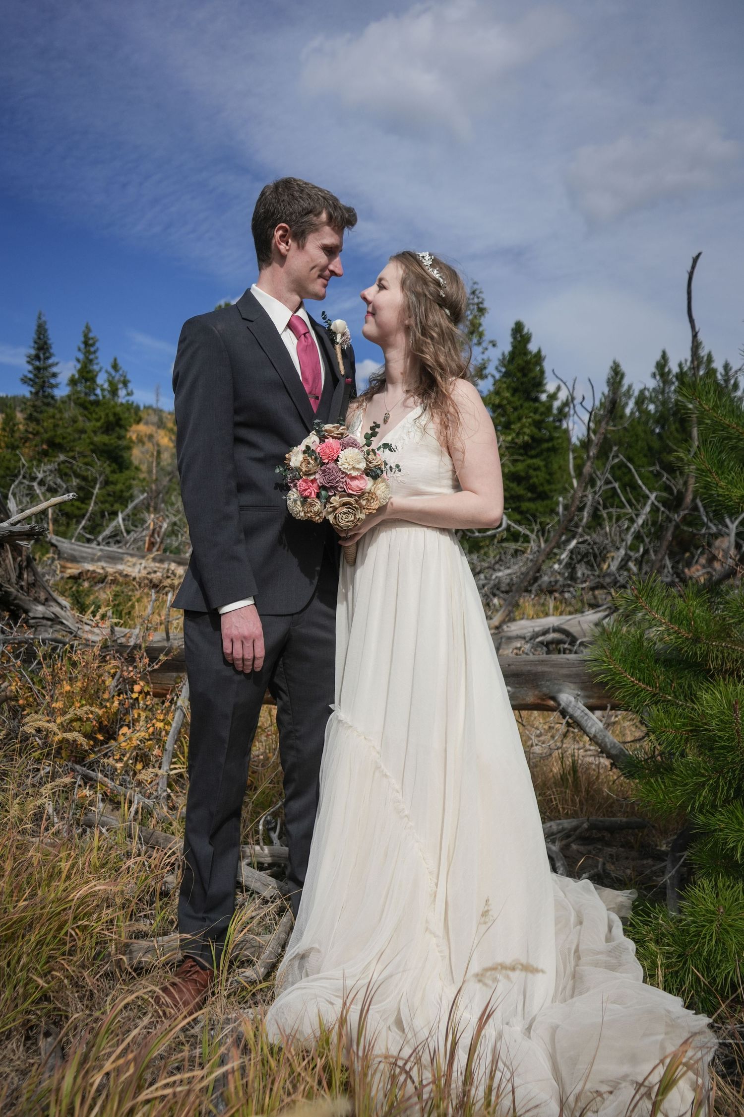 A wedding couple poses happily in formal attire against a mountain landscape with pine trees.