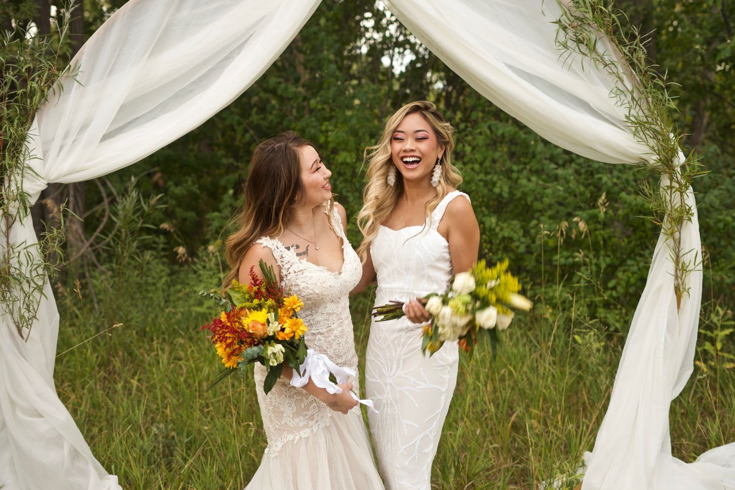 Two smiling brides in white wedding dresses holding colorful bouquets under a draped white fabric arch.