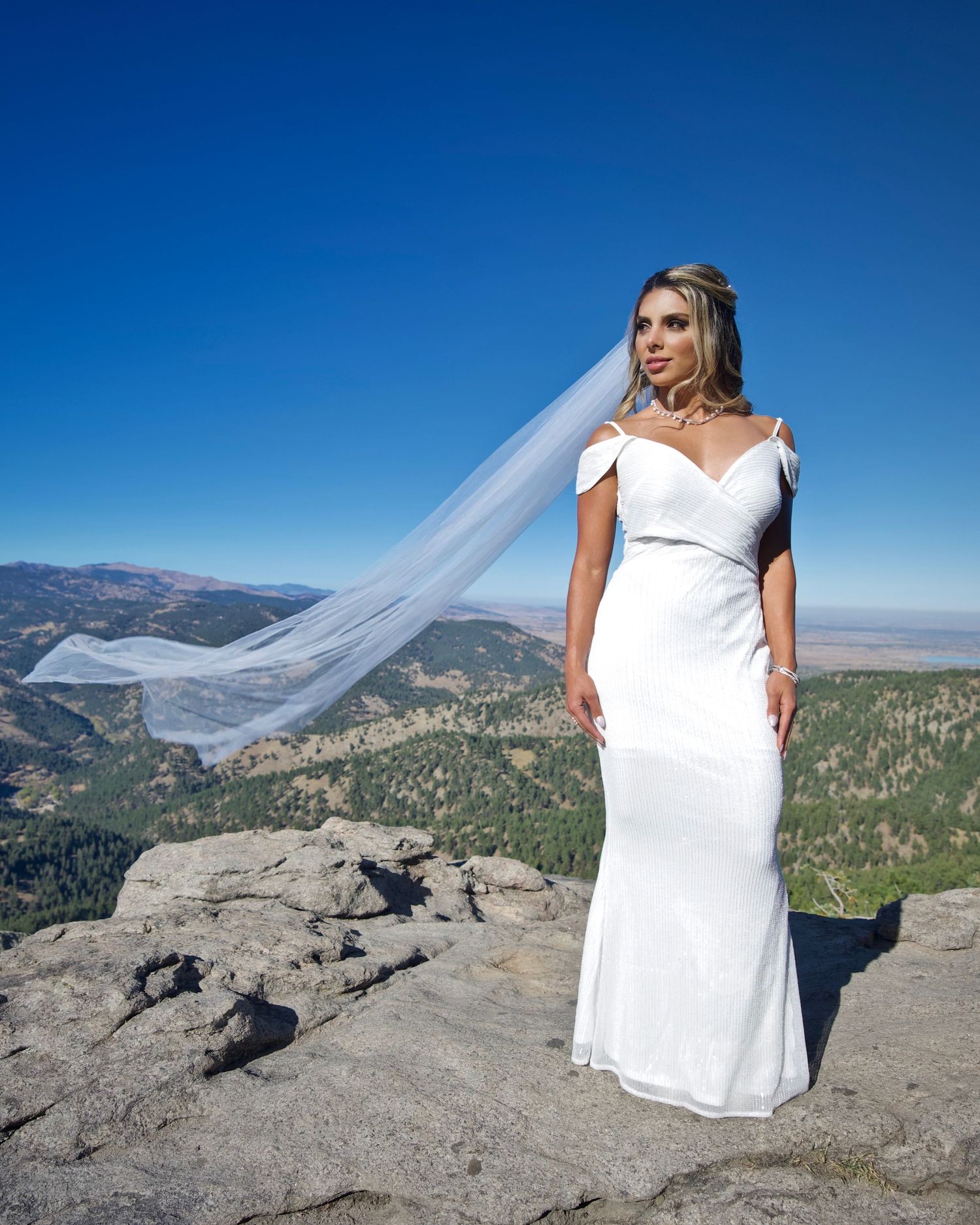A bride in a white wedding dress with flowing veil poses dramatically on a mountain summit.