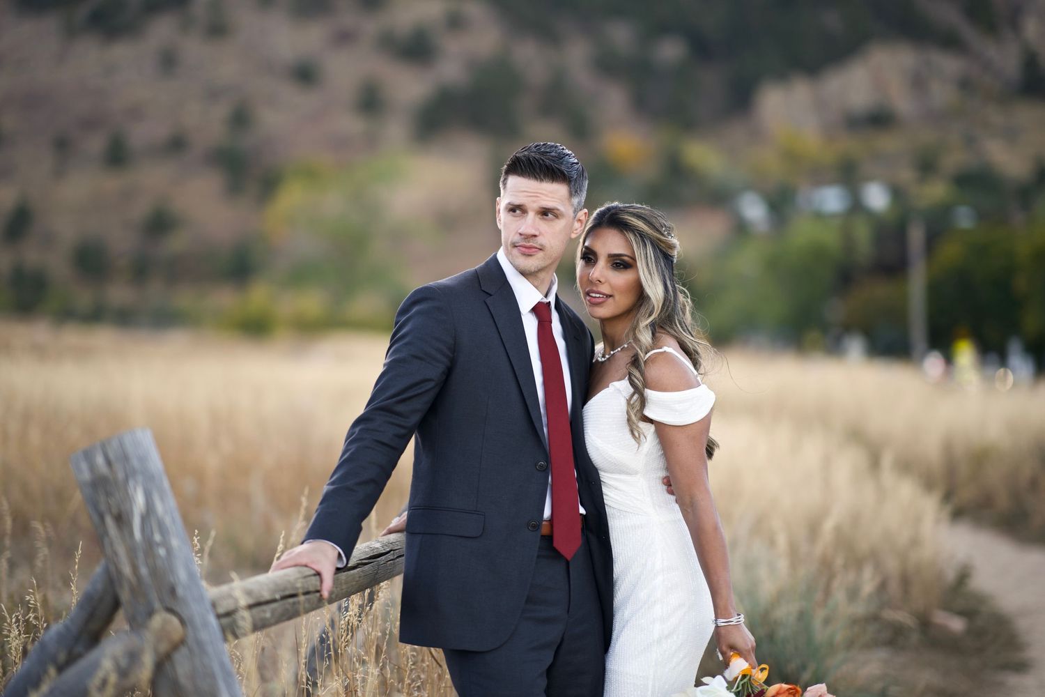 A wedding couple poses together in formal attire against a rustic mountain landscape.
