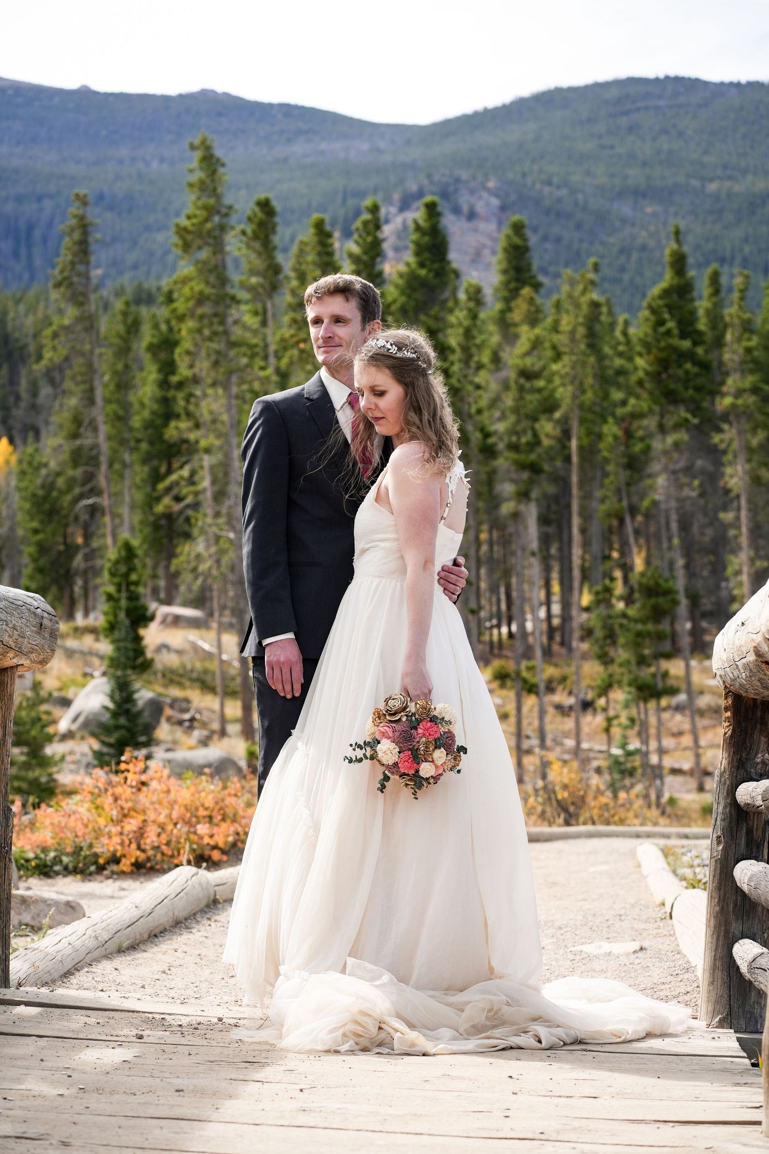 Newlyweds stand on a wooden bridge with mountain views and fall colors in the background.