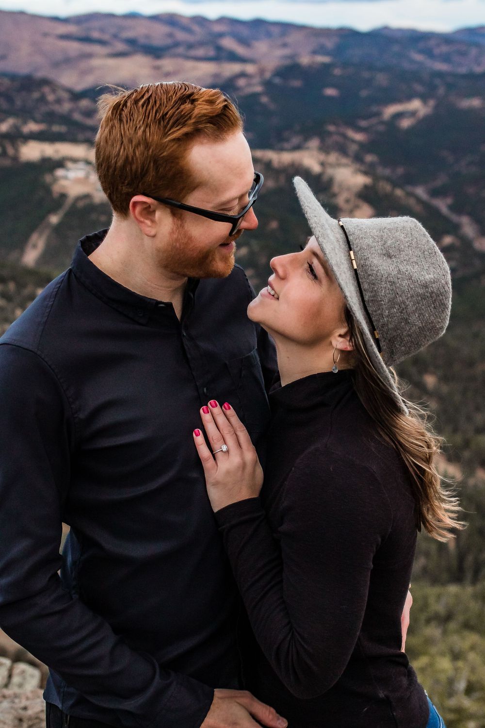 Intimate couple in dark clothing and gray hat share close moment against mountain backdrop. Colorado engagement photography.