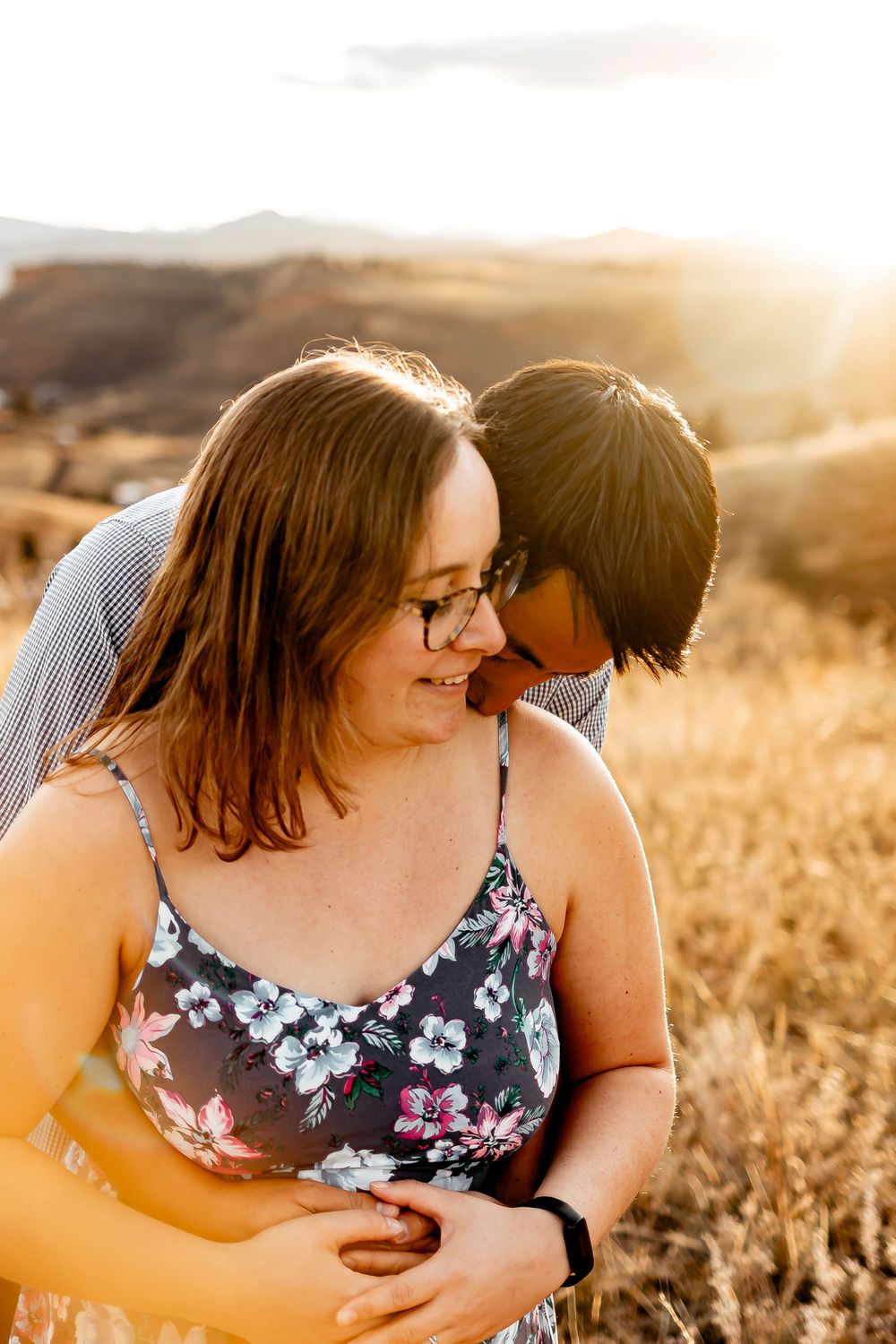 Couple embracing and laughing together in golden sunlit field with mountains in background. Fort Collins Engagement Photography.