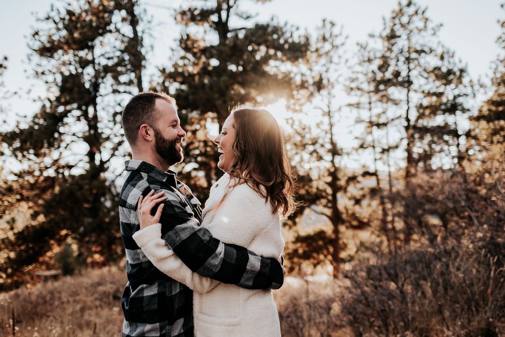 Couple in casual winter attire embraces while laughing together in sunlit forest setting. Colorado engagement photography.