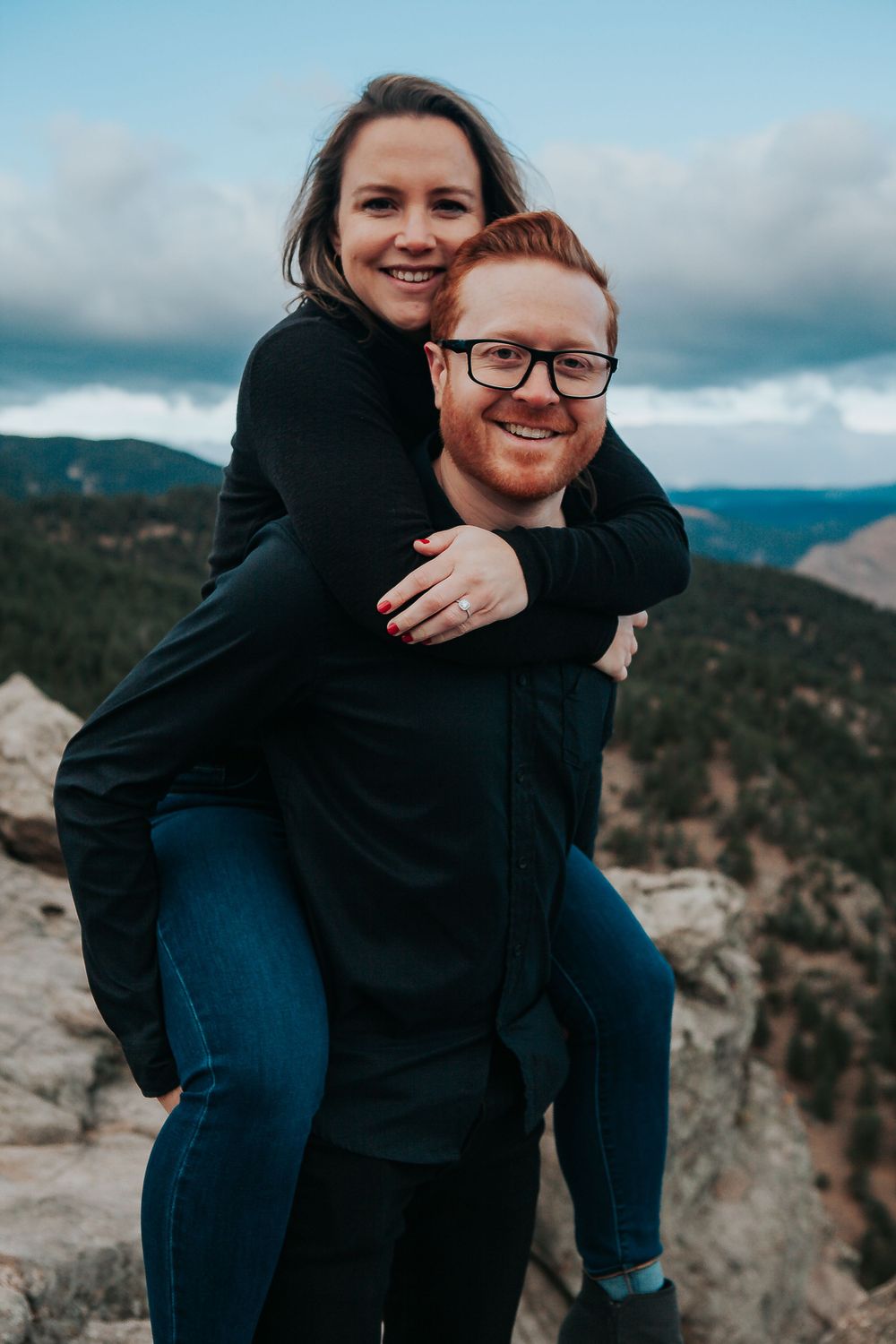 Happy couple enjoys piggyback ride on mountain summit with scenic valley views behind them. Colorado engagement photography.