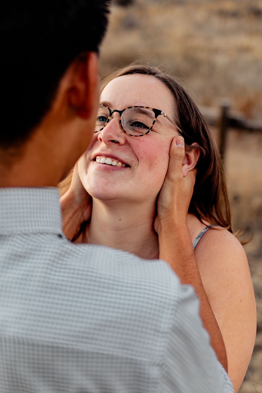 Person wearing glasses smiles warmly as their partner touches their face in a golden field. Fort Collins Engagement Photography.