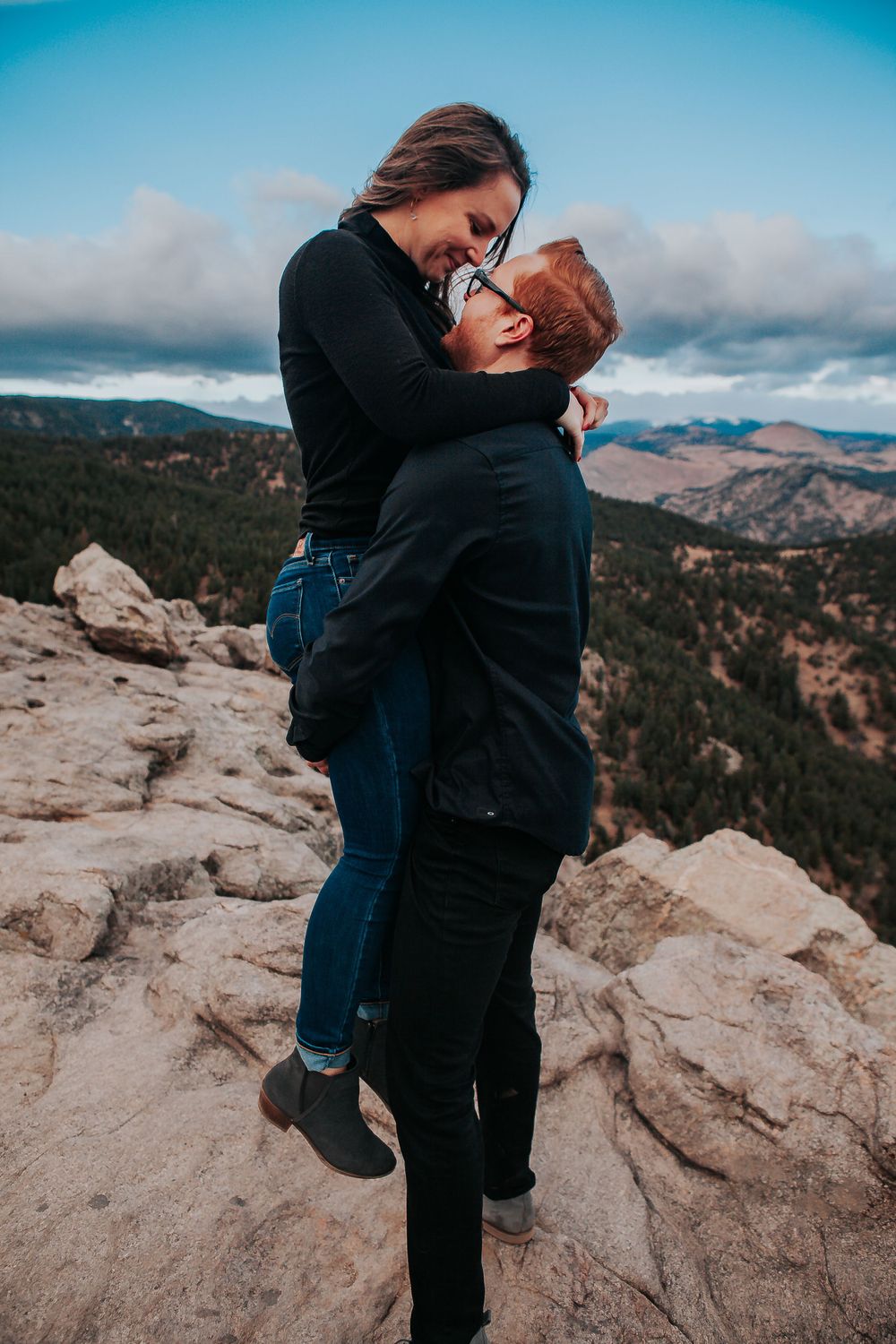 Couple shares romantic moment on mountain cliff overlooking scenic valley landscape. Colorado engagement photography.