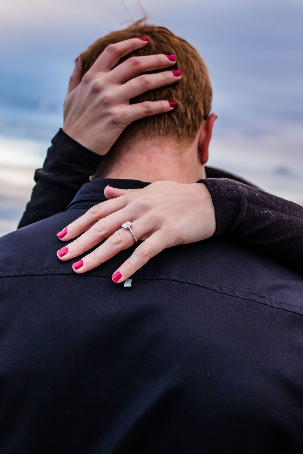 Close-up of hands with bright pink nails and engagement ring embracing from behind. Colorado engagement photography.