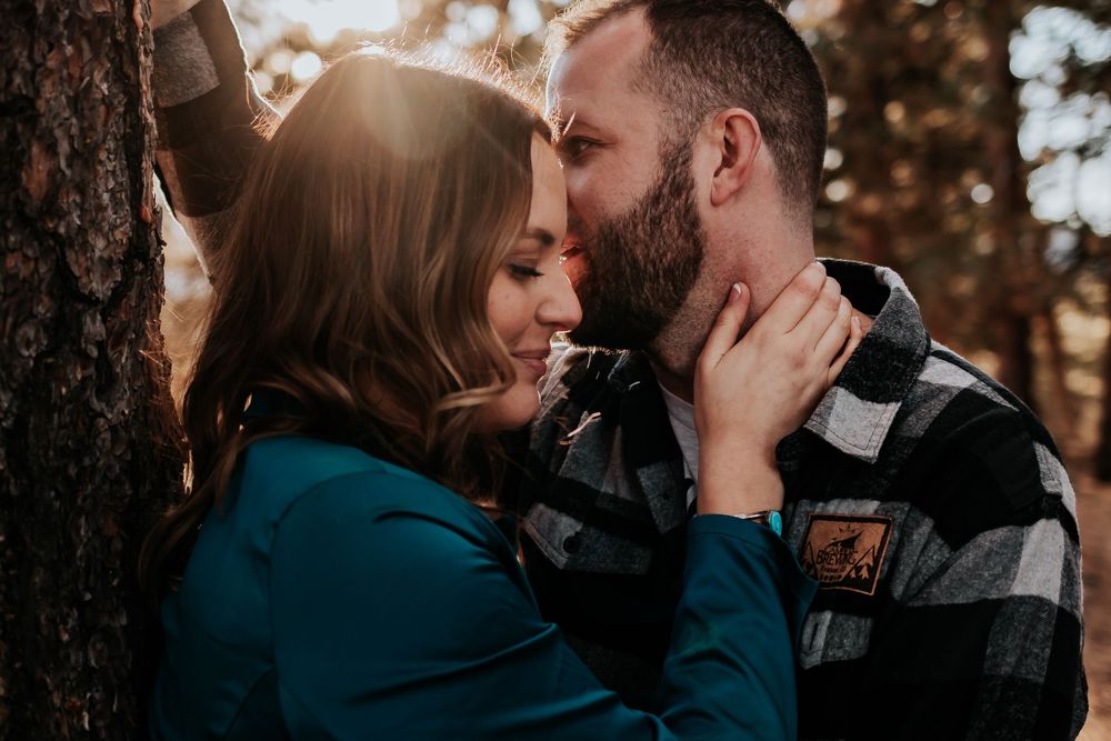 Intimate couple shares tender moment against tree trunk in autumn forest with sun flare. Colorado engagement photography.