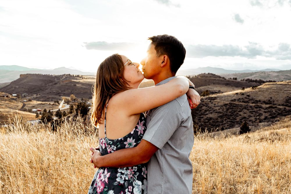 Couple sharing a romantic kiss against a backdrop of golden hills and mountains at sunset. Fort Collins Engagement Photography.