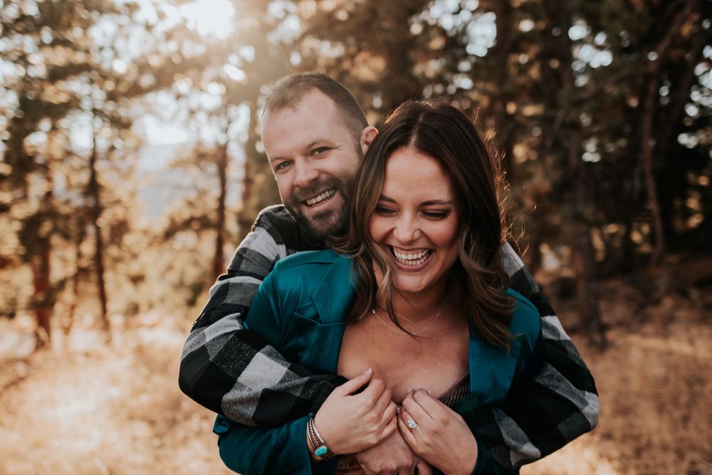 A joyful couple embraces outdoors in a forest during golden hour wearing plaid and teal clothing. Colorado engagement photography.
