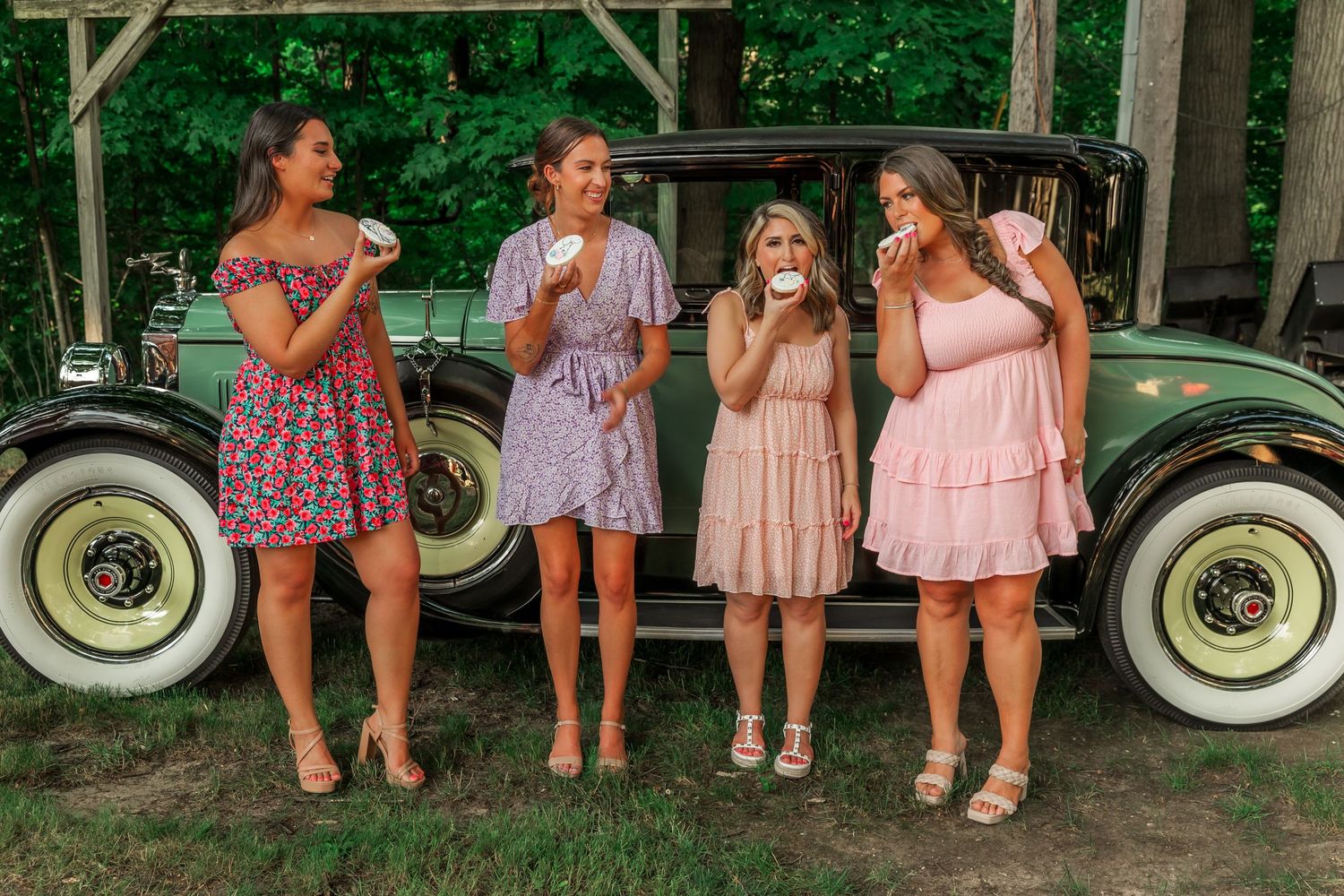 Group poses in coordinated summer dresses beside vintage car at outdoor celebration.
