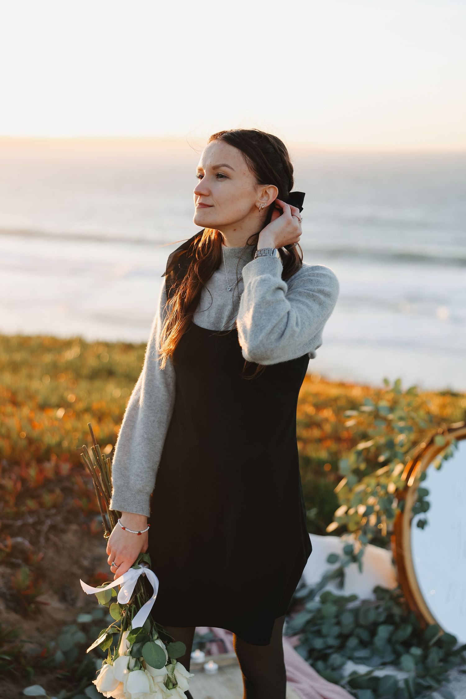 Woman in black dress and gray sweater holding flowers against ocean backdrop.