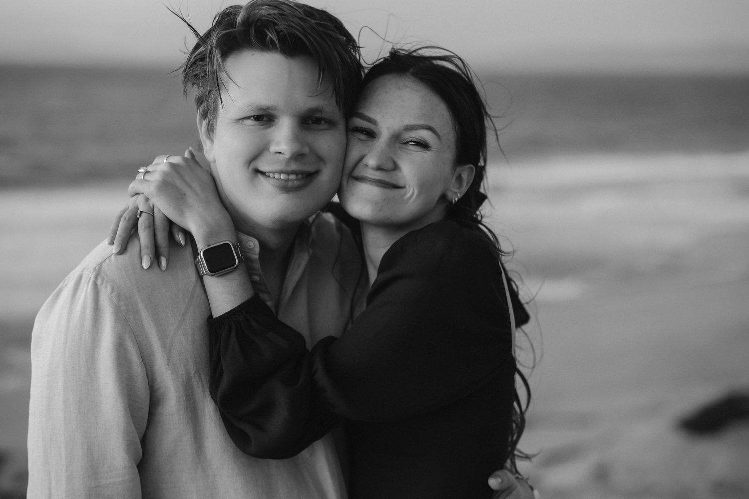 A black and white portrait of a couple embracing on a windswept beach with waves rolling in behind them.