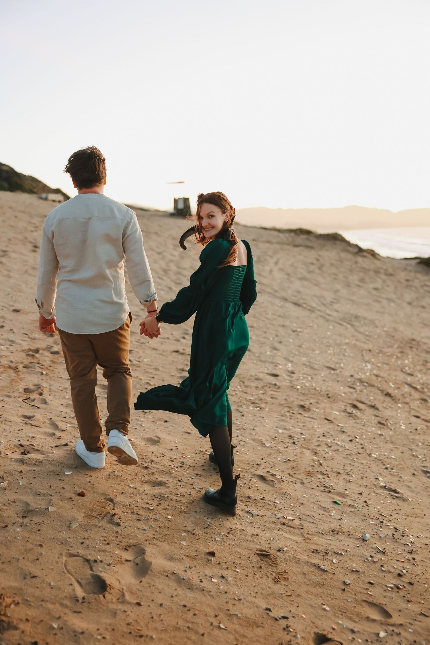 Couple in formal wear walks hand in hand along coastal dunes at golden hour.