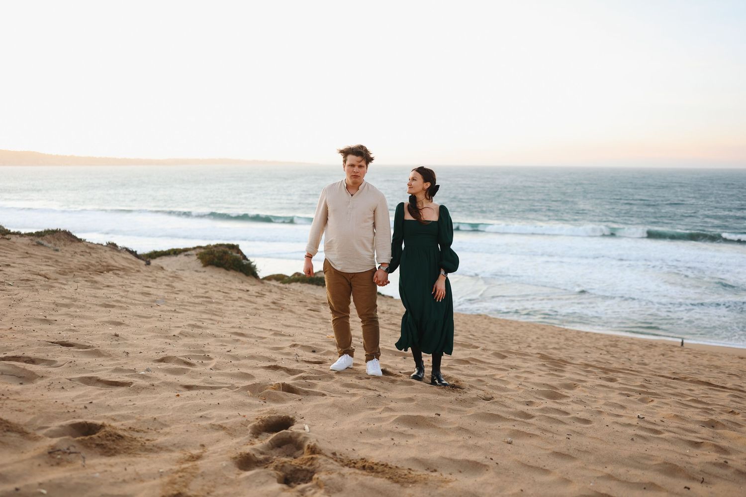 A couple takes a leisurely stroll together on a sandy beach with waves crashing behind them.