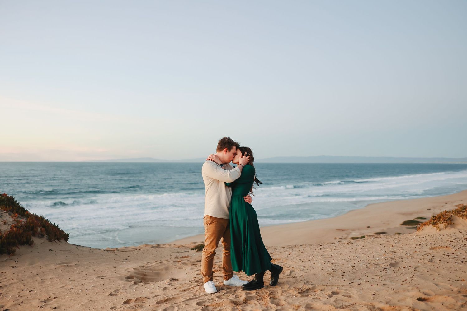 A serene beach engagement photo with two people in casual attire sharing an intimate moment by the ocean.
