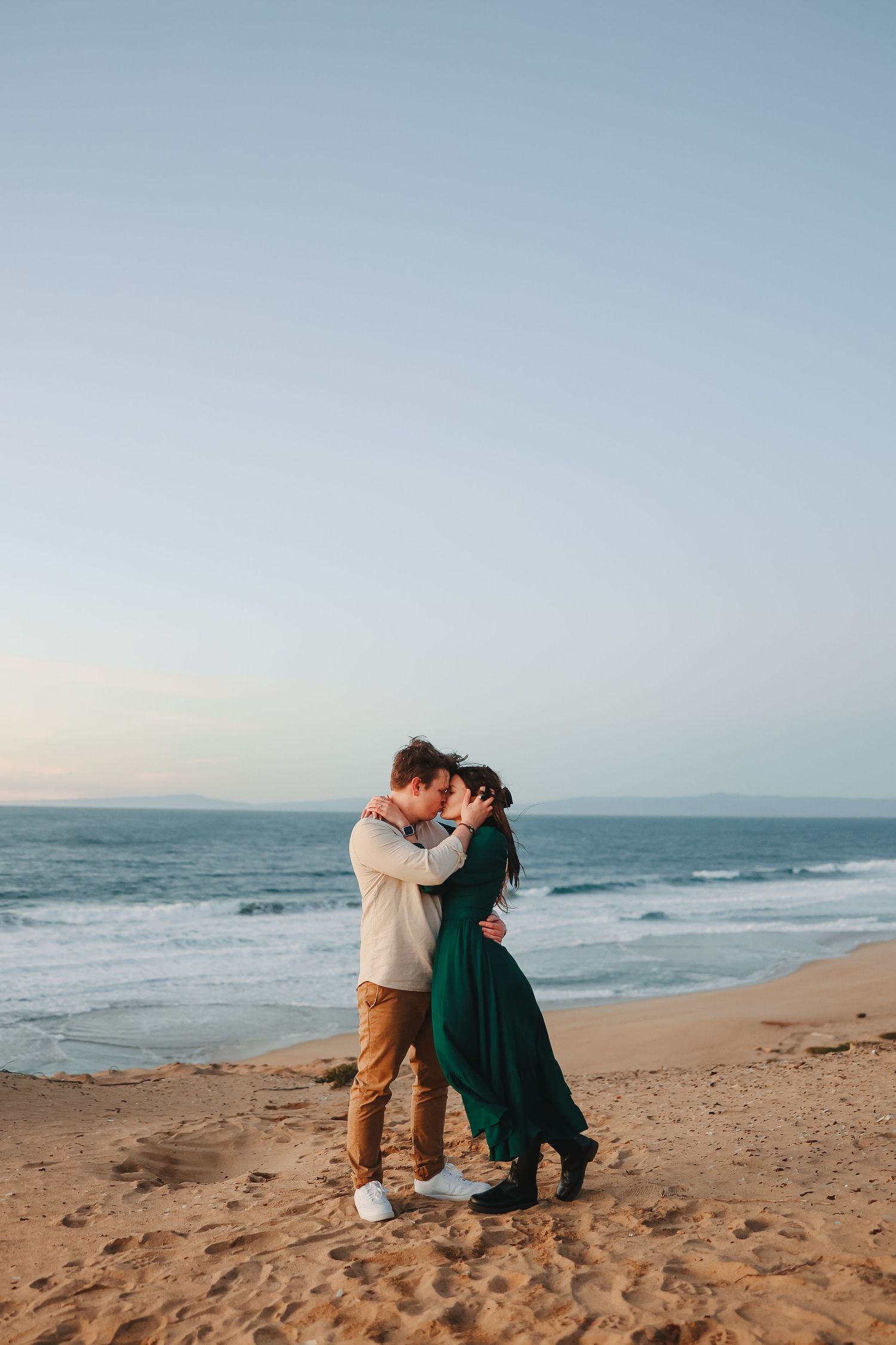 A romantic seaside portrait of two people embracing on sandy beach during golden hour.