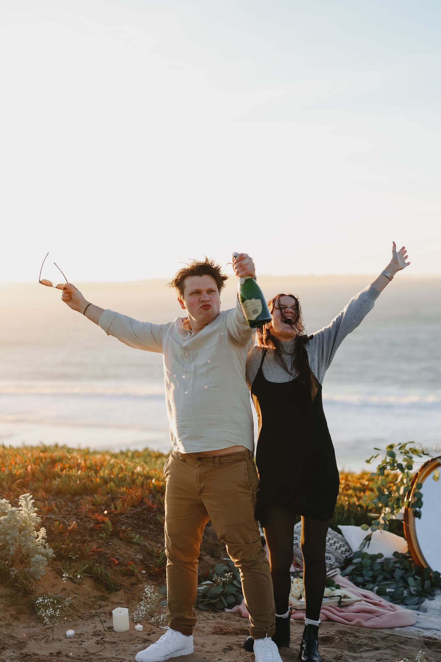 Couple celebrating with drinks at sunset on a coastal cliff with ocean views in background.