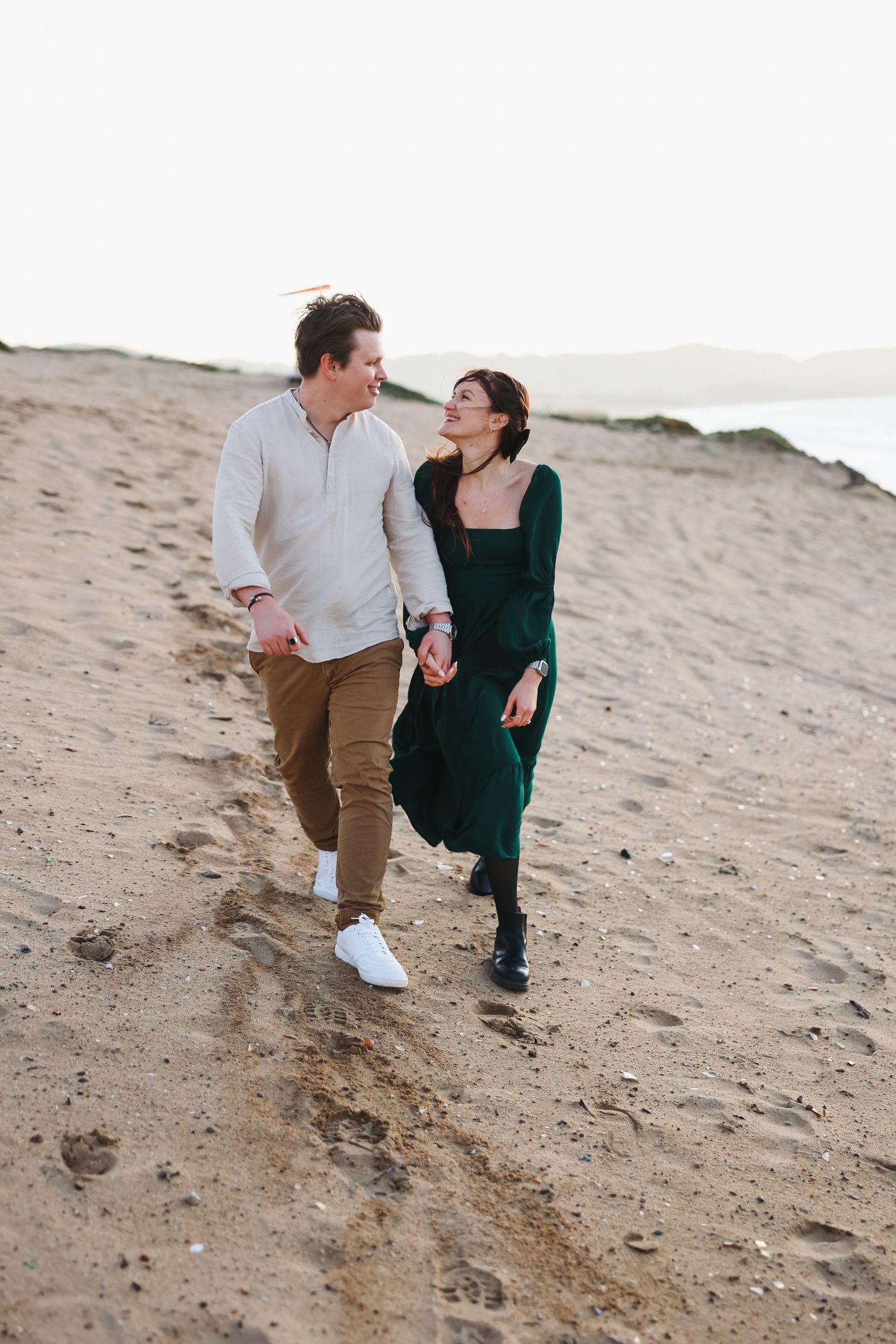 A couple walks hand in hand on a sandy beach path wearing casual attire.