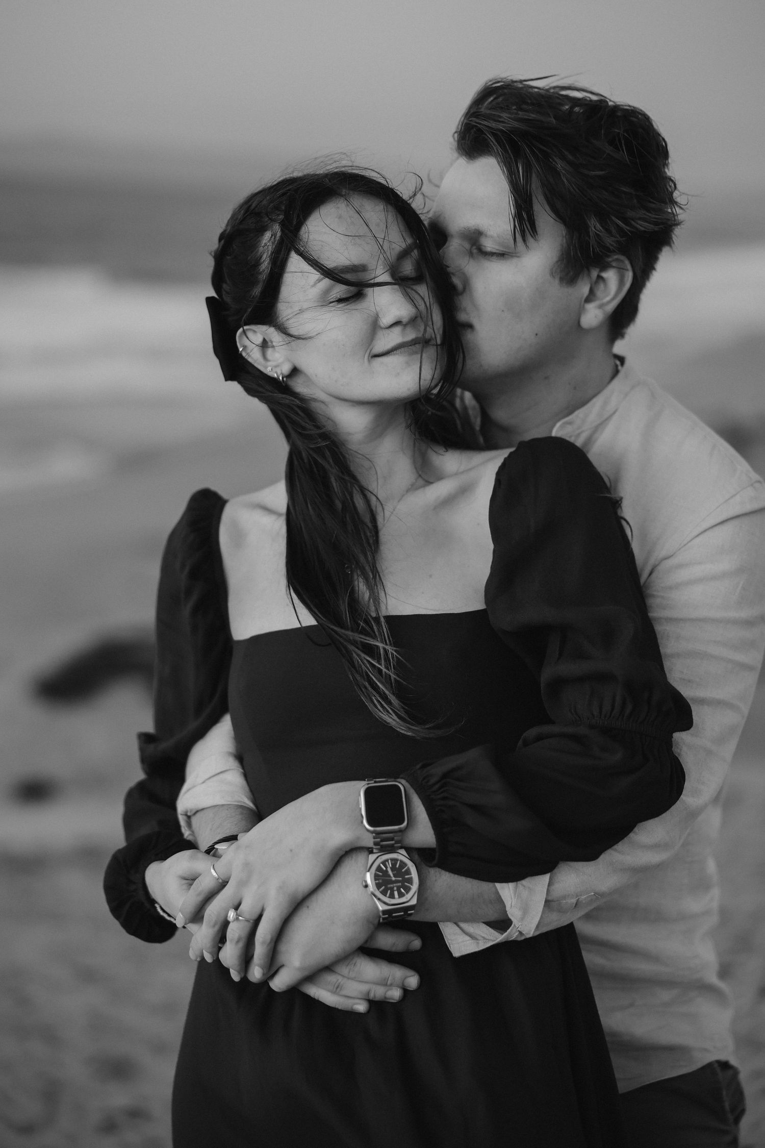 Black and white photo of an intimate beach embrace, showing smartwatches and a flowing dark dress.