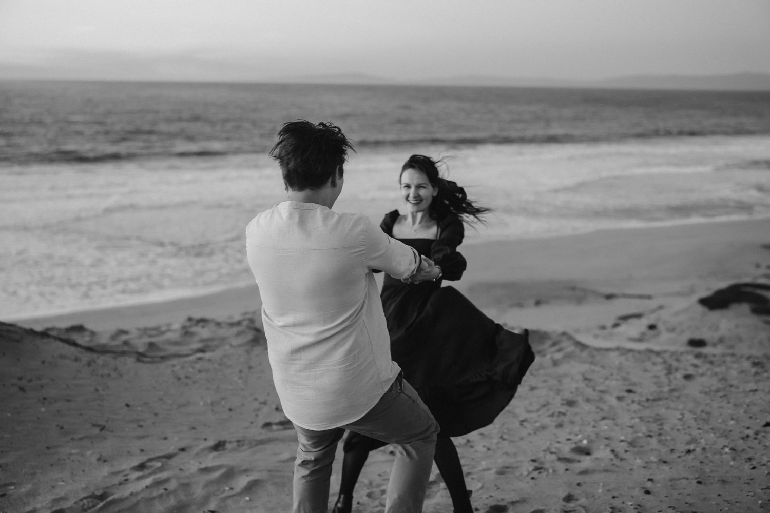 A black and white photo shows a couple dancing joyfully by the ocean.