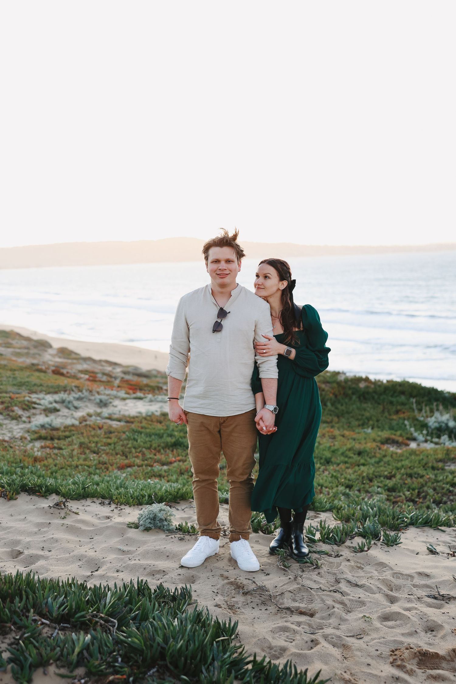 A couple in casual attire stands together on a coastal path during sunset.