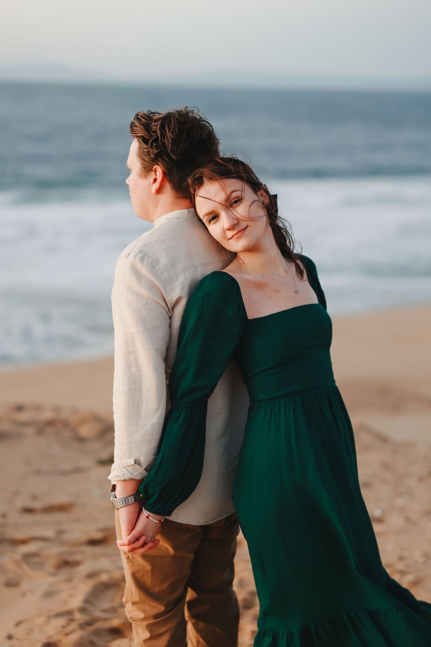A couple in contrasting outfits poses romantically against an ocean backdrop at sunset.