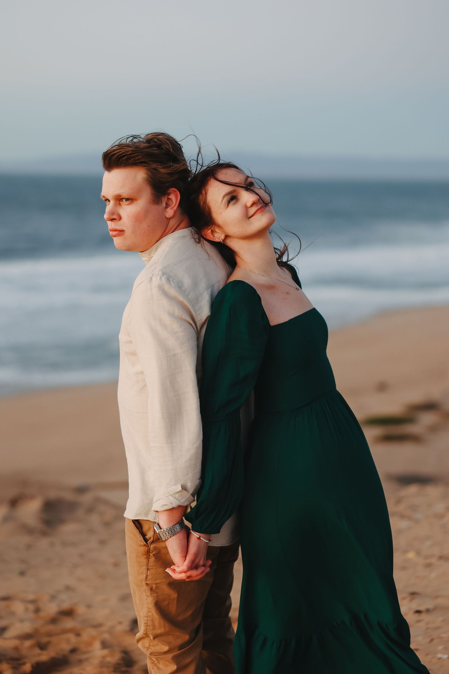 A romantic couple standing back-to-back on a beach at sunset, with one wearing a cream sweater and the other in emerald dress.