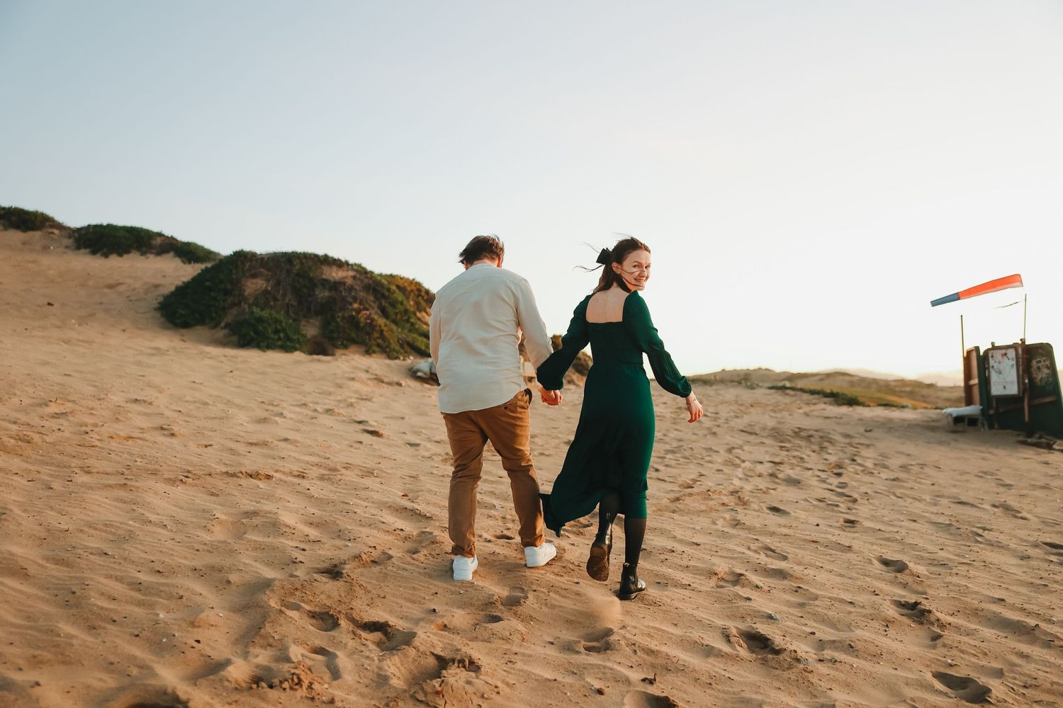 Couple walking together on sandy dunes with emerald dress flowing in coastal breeze.