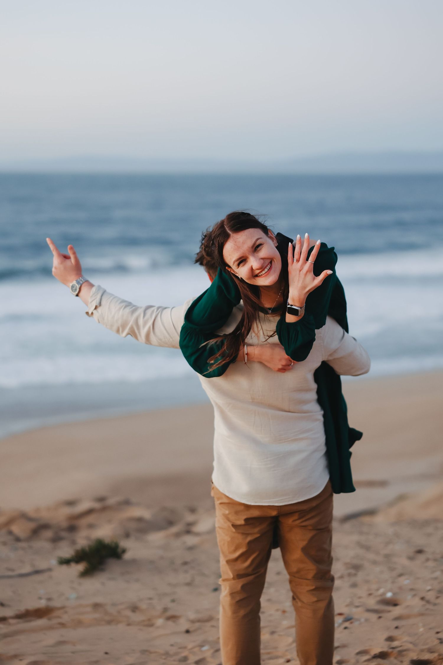 A playful person in a cream sweater and brown pants poses joyfully against an ocean backdrop at sunset.