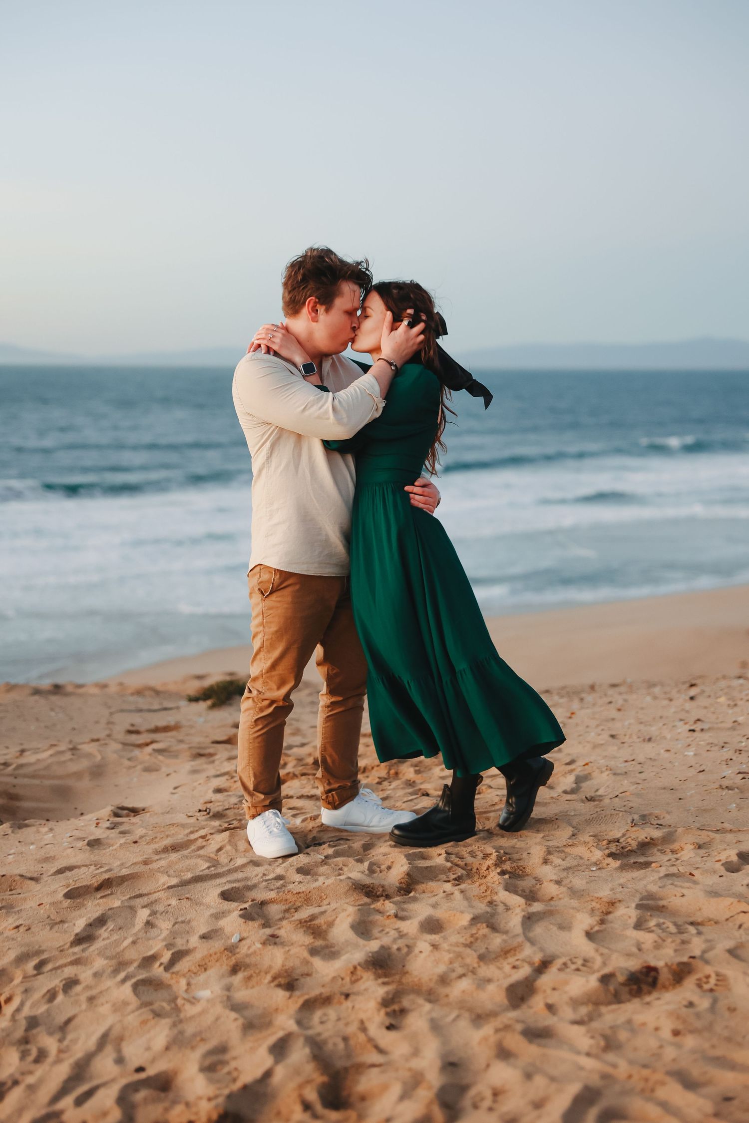 A couple sharing a romantic moment on a beach, one in beige sweater and other in flowing green dress.