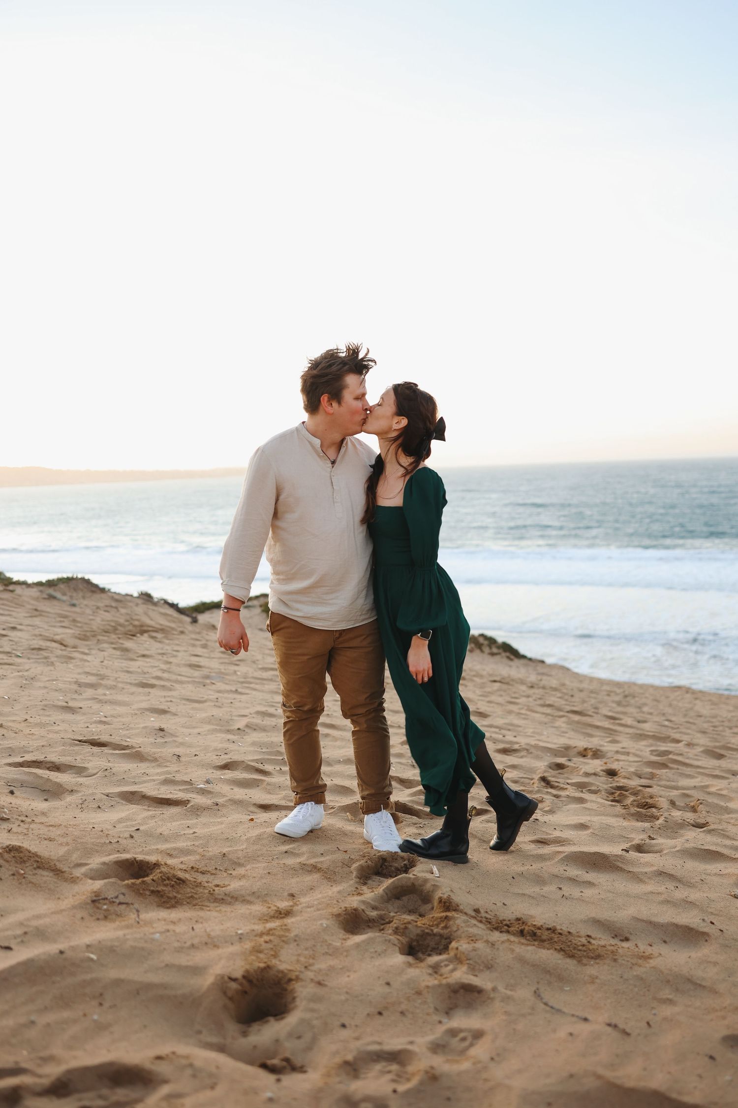 A couple in casual wear embraces while standing on a beach with ocean waves in the background.