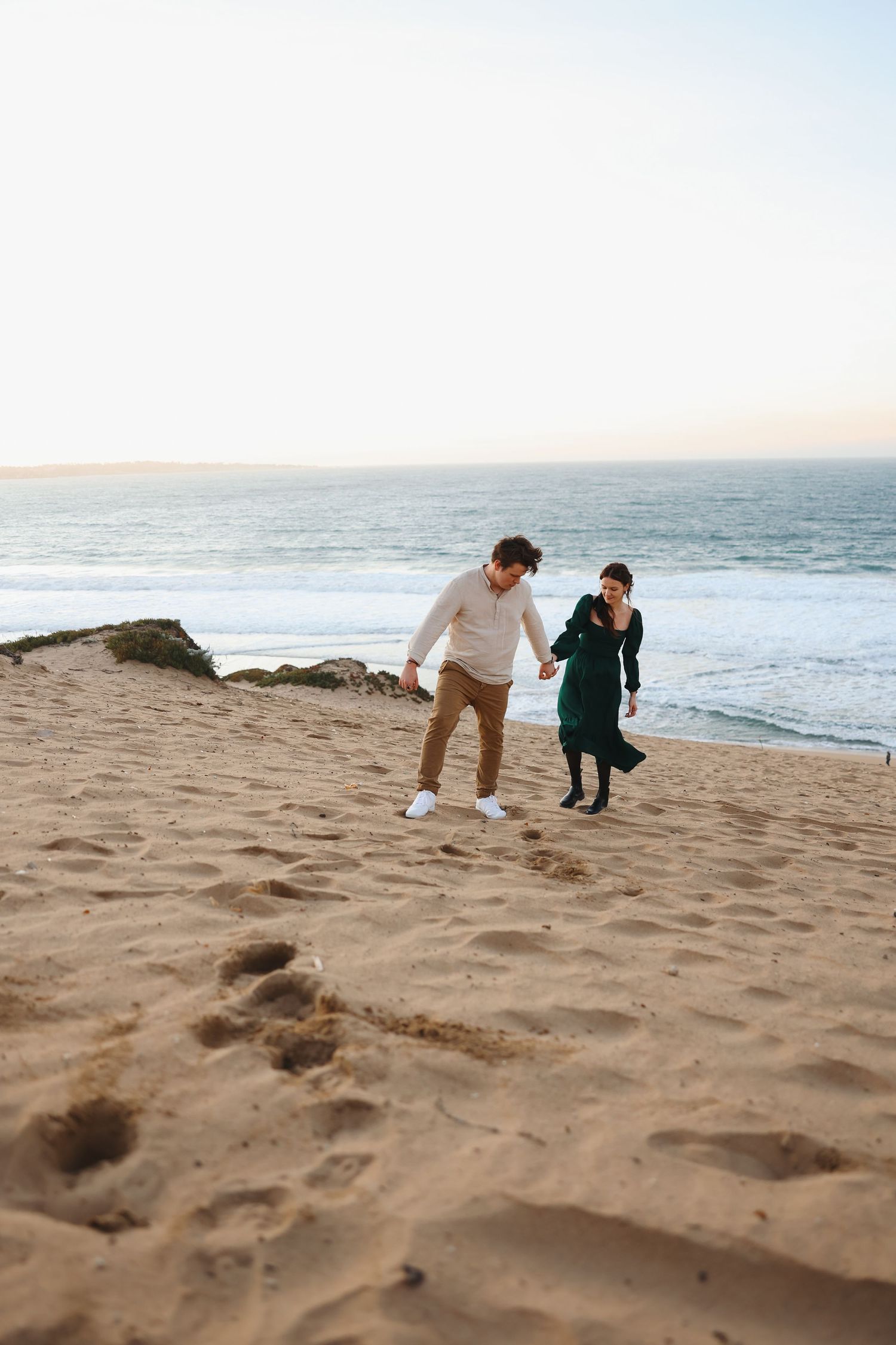 A couple runs down a sandy beach hill together with the ocean visible in the background.