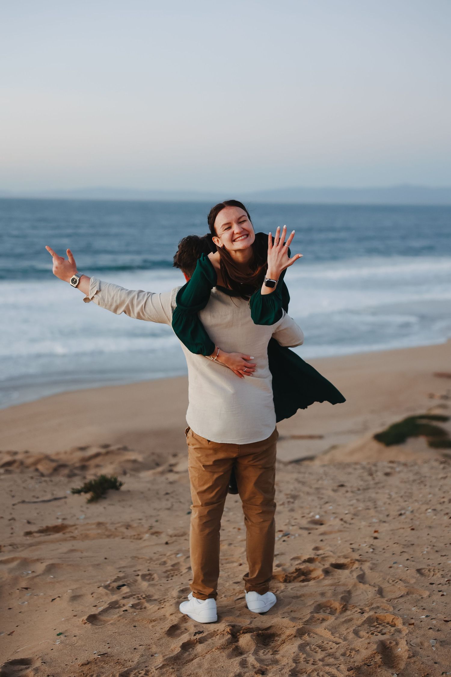 Playful engagement photo with couple striking a fun pose on sandy beach at sunset.
