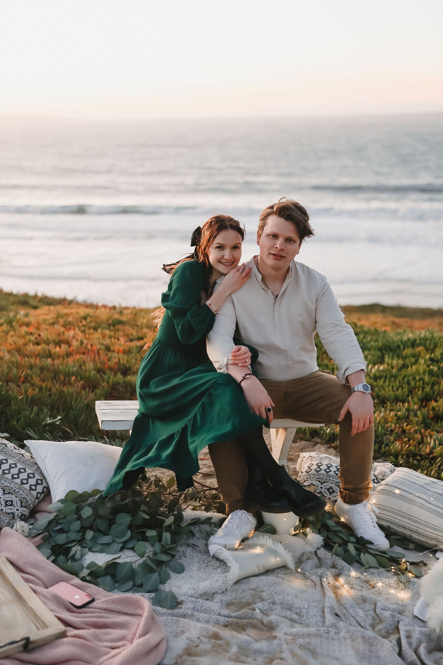 A romantic sunset picnic setup on a coastal cliff with blankets and candles overlooking the ocean.