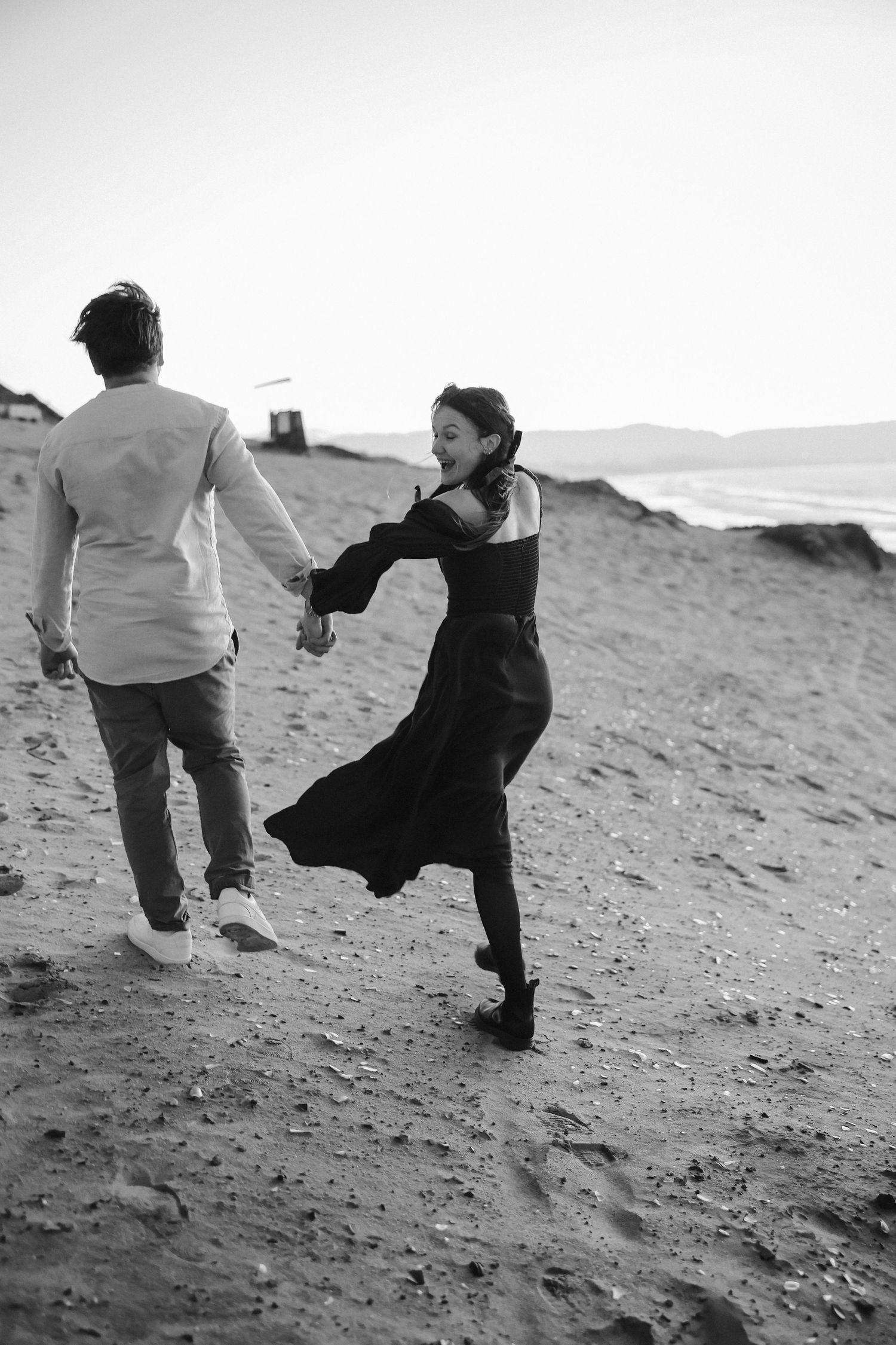 Black and white photo of couple dancing joyfully on windswept beach dunes.